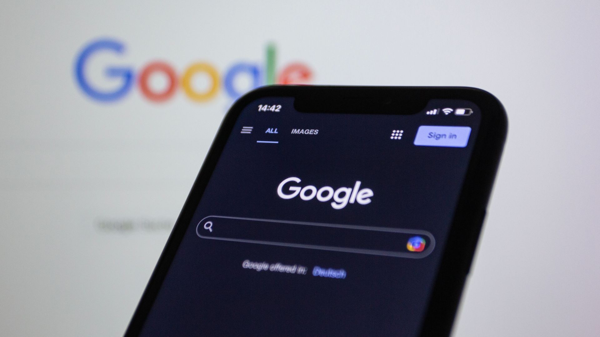 Smartphone mit geöffneter Google-Suche, dahinter unscharf das Google-Logo auf einem Bildschirm